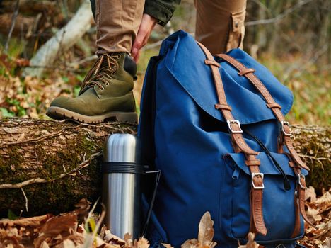 Female hiker wears boots outdoors in autumn forest, near thermos and backpack. View of legs. Hiking and leisure theme