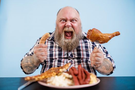 Emotional bearded obese man holds sausage and smoked chicken leg sitting at table with rich food on light blue background in studio