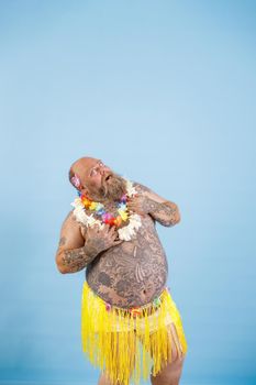 Surprised middle aged plump man in decorative yellow grass skirt and flowers garland looks up posing on light blue background in studio