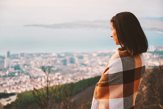 Young woman wrapped in plaid looking at the city from hill in autumn. Concept of tranquil life from the urban bustle