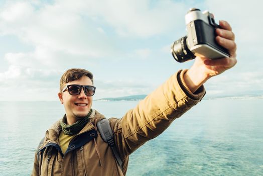 Traveler happy young man takes photographs self portrait with old photo camera on coastline on background of sea