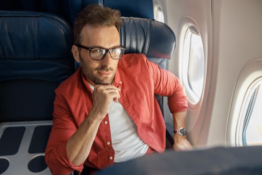 Attractive caucasian businessman in casual wear and glasses looking thoughtful, sitting on the plane near the window. Relax, travel, vacation, transportation concept