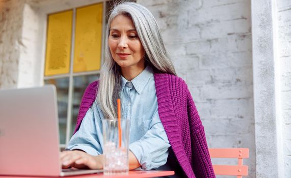 Mature Asian businesswoman with loose hair types on laptop keyboard sitting at table on outdoors cafe terrace on autumn day