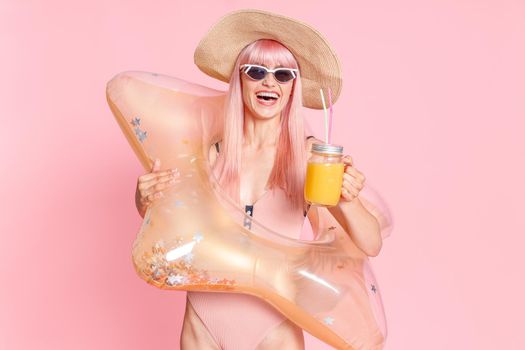 Joyful woman with pink hair in swimsuit and sunglasses with tropical cocktail and inflatable star for swimming posing isolated over pink background. Summer vacation, fun, party concept