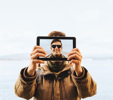 Hiker young man in sunglasses doing a self-portrait with him digital tablet on background of sea