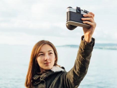 Smiling young brunette woman takes photographs selfie portrait with old photo camera on coastline on background of sea