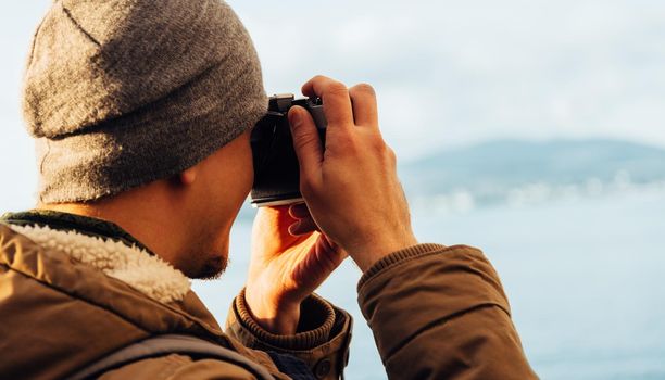Traveler hipster guy takes photographs with vintage photo camera on coastline near the sea