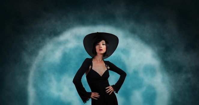 Attractive fashionable witch in black hat and dress on background of full moon. Halloween theme