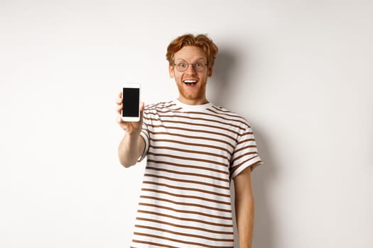 Technology and e-commerce concept. Happy young redhead man in glasses showing blank smartphone screen, looking at camera amazed, standing over white background.