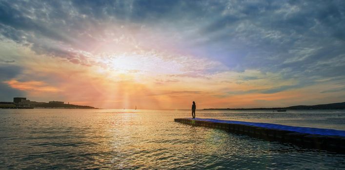 Woman standing on pier near the sea and looking at sunset, tranquil scene. Beautiful seascape. Image with sunlight effect