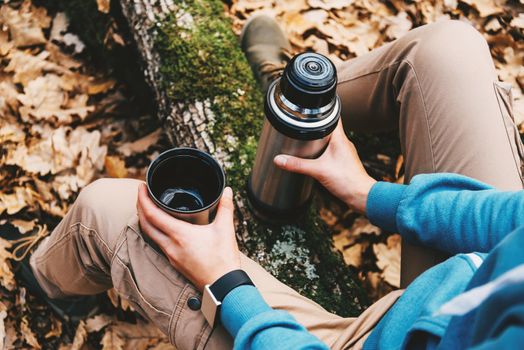Unrecognizable traveler woman holding a cup of tea or coffee and thermos in autumn forest. Hiking and leisure theme