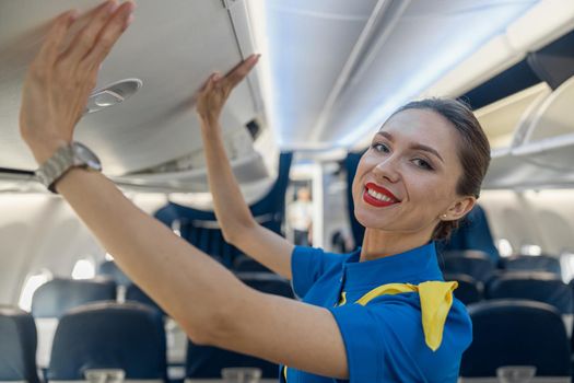 Portrait of pretty stewardess in blue uniform smiling at camera while closing hand luggage compartment, staying on the aisle inside the plane. Transportation, occupation concept