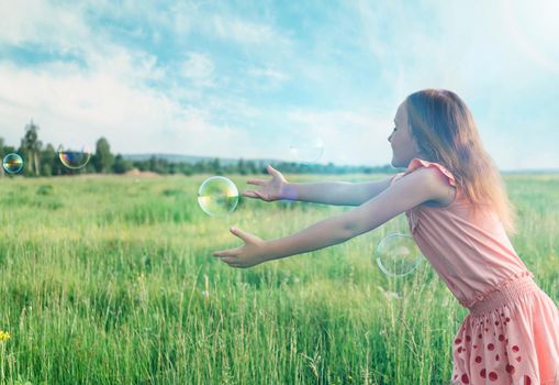 Cheerful little girl playing among soap bubbles on summer meadow outdoor. Concept of happiness and carefree