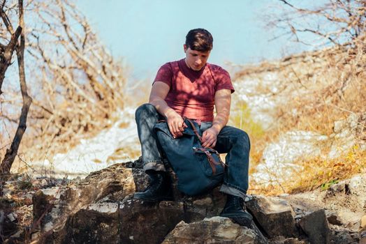 Young hiker man opening his backpack on nature on a halt resting. Hiking and recreation theme