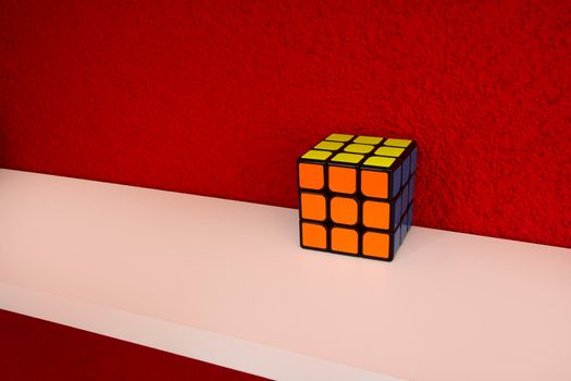 Solved puzzle speed cube on white wooden shelf near red colored wall, colorful rubiks cube with copy space mind challanging retro design