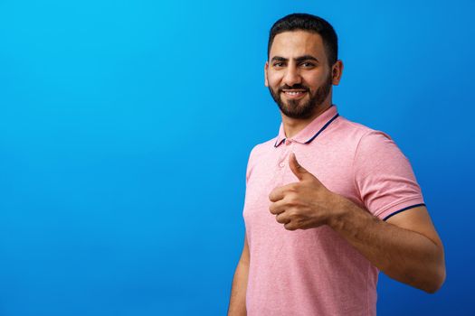 Happy beard young man smiling with thumbs up against blue background, close up