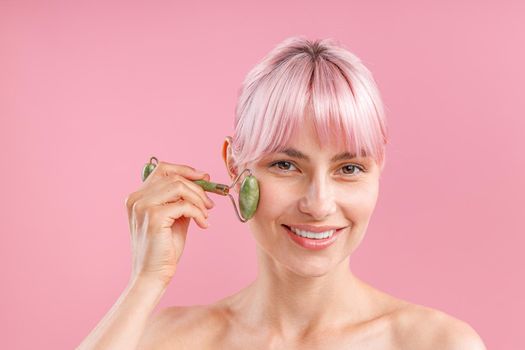 Cheerful naked woman with pink hair smiling at camera while using natural jade facial roller for skin care isolated over pink background. Face massage and beauty trends concept