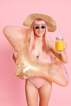 Cheerful woman with pink hair in swimsuit and sunglasses with tropical cocktail and inflatable star for swimming posing isolated over pink background. Summer vacation, fun, party concept