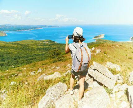 Hiker young woman with backpack takes photographs beautiful sea and standing on mountain, rear view