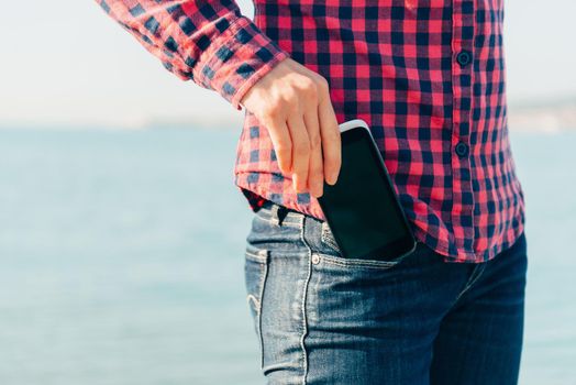 Woman takes out mobile phone of her pocket of jeans on beach near the sea to make self-portrait or to photograph the sea