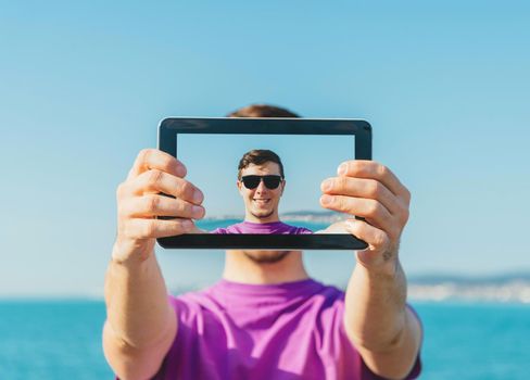 Young smiling handsome man doing a self-portrait with him digital tablet on background of sea