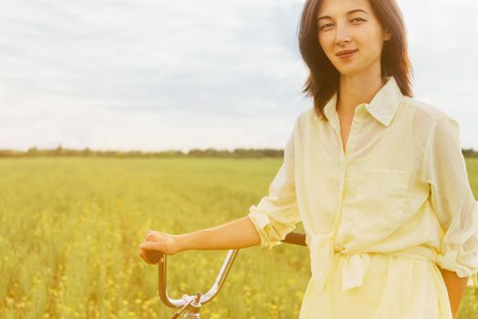 Beautiful young woman walking with bicycle on summer meadow at sunny day. Image with sunlight effect
