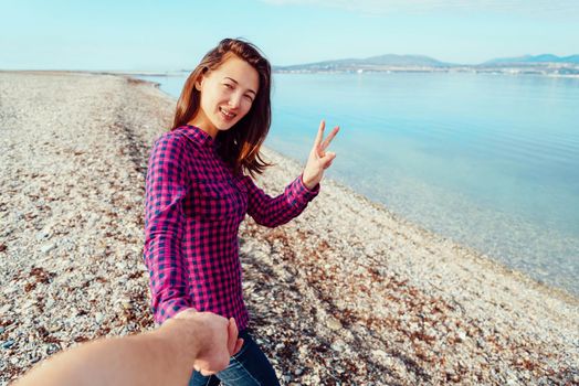 Couple in love. Smiling young woman holding man's hand and showing peace sign on beach near the sea