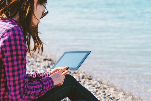 Young woman sitting on beach near the sea and working on digital tablet. Focus on face