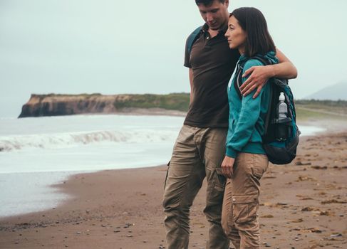 Traveler couple in love with backpacks walking on beach near the sea in summer. Man embracing a woman