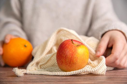 Female hand with environmentally friendly net bag for shopping groceries with Fresh fruit, Apples and oranges, eco friendly net on wooden table close up