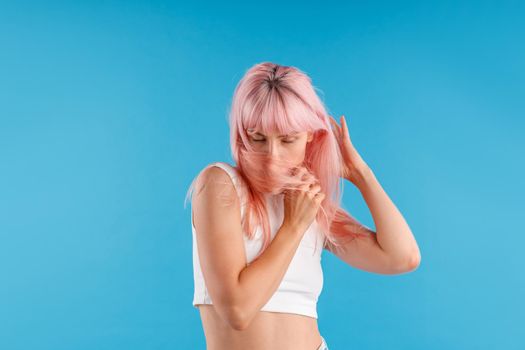 Attractive female model in white shirt looking down, hiding her face with pink hair while posing isolated over blue studio background. Beauty, hair care concept