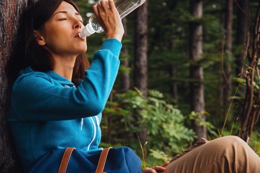Hiker young woman rests near a tree and drinks water in summer forest