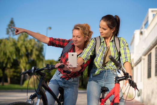 Two pretty young caucasian girls searching the way on a bicycles with smartphone. Best friends enjoying a day on bikes. Sunny summer evening.