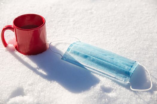 Red mug with hot coffee,tea or chocolate milk and protective blue medical mask for coronavirus Covid-19 pandemic. lies in the snow, Winter,virus,drink and health concept background beauty