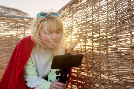 Outdoor sunset portrait of female teenager recording video on smartphone, rattan, sky background. Video call, stream, blog, vlog, telecommunications technologies, youth concept, copy space