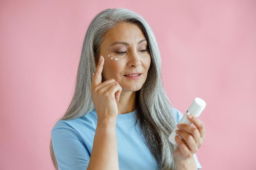 Pretty middle aged Asian woman applies cream under eyes holding bottle on pink background in studio. Mature beauty lifestyle