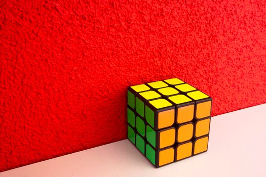 Solved puzzle speed cube on white wooden shelf near red colored wall, colorful rubiks cube with copy space mind challanging retro design