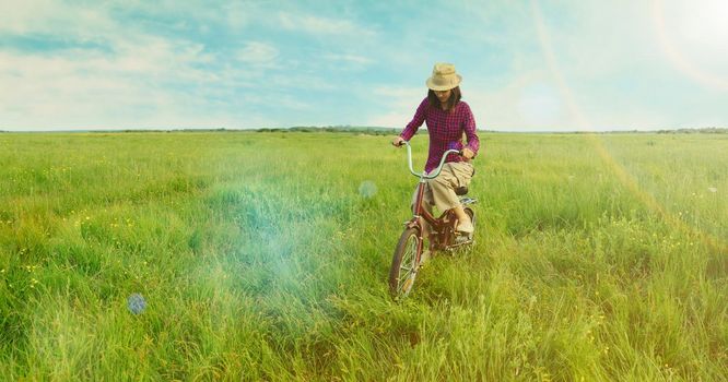 Hipster young woman cycling on green meadow in summer. Image with sunlight effect