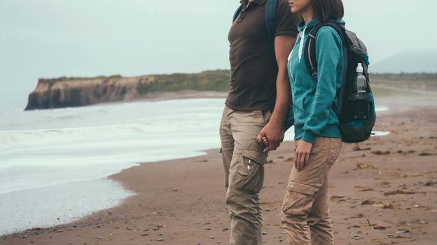 Traveler couple in love with backpacks walking on coastline near the sea in summer and holding each other hand