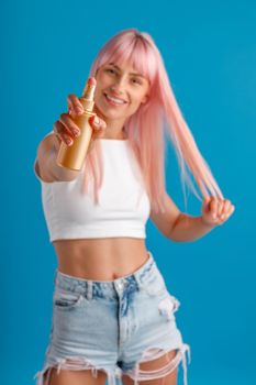 Cheerful young woman with pink hair smiling and showing moisturizing spray to camera while standing isolated over blue studio background. Beauty products, hair care concept