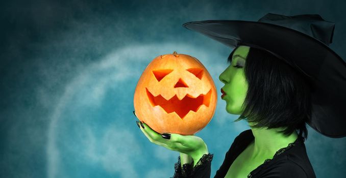 Witch with green skin kisses a Halloween pumpkin at midnight