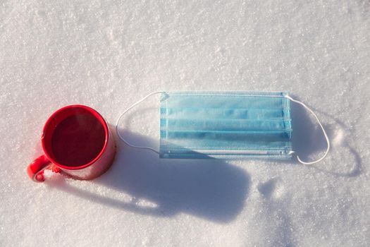 Red mug with hot coffee,tea or chocolate milk and protective blue medical mask for coronavirus Covid-19 pandemic. lies in the snow, Winter,virus,drink and health concept background beauty