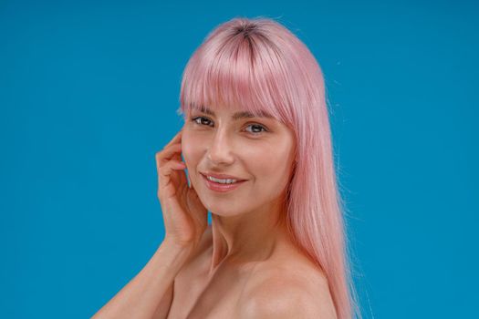 Portrait of beautiful young woman with pink hair and perfect skin smiling at camera, posing isolated over blue studio background. Beauty, skin care concept