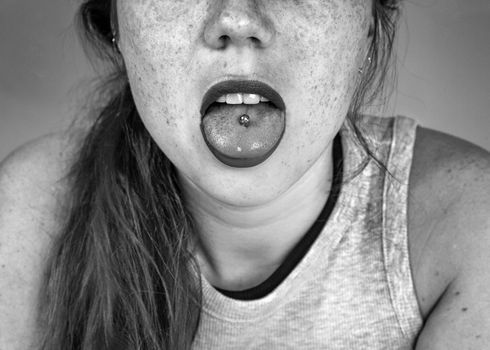 Close up portrait of young woman sticking out pierced tongue, showing her tongue piercing with black and white closeup