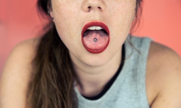 Close up portrait of young woman with freckles sticking out pierced tongue, showing her tongue piercing with pink background front view