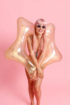 Smiling young woman with pink hair in swimsuit and sunglasses with inflatable star for swimming posing isolated over pink background. Summer vacation, fun concept