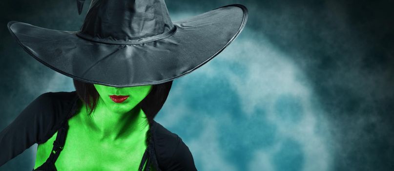 Beautiful fashionable witch with green skin in black hat and dress on background of full moon. Halloween theme