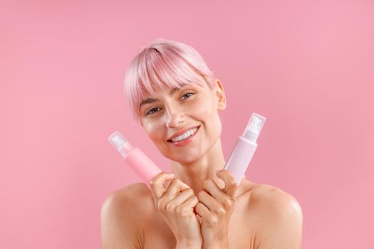 Portrait of lovely young woman with pink hair holding two plastic bottles with beauty products, posing isolated over pink background. Beauty, spa, body care concept