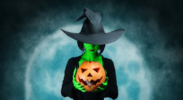 Mystery witch with green skin holds Halloween pumpkin with gray rat at midnight