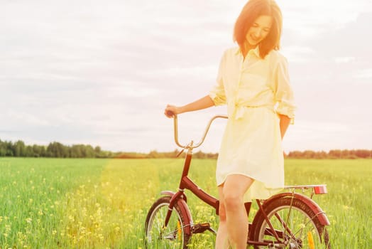 Beautiful young woman in dress walking with bicycle on summer meadow at sunny day. Image with sunlight effect